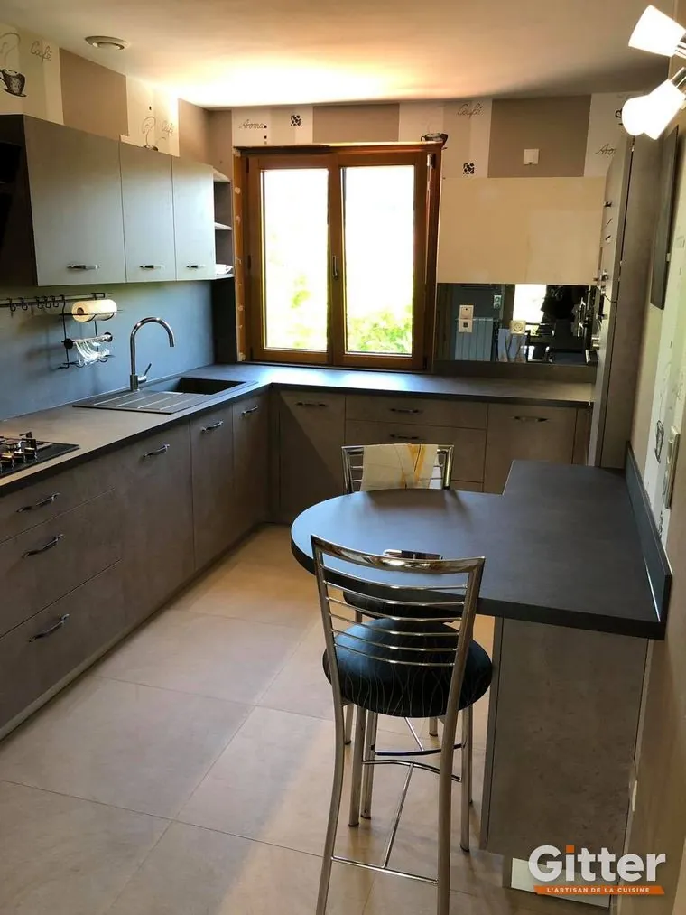 Cuisine en L, façades mates gris taupe, plan de travail foncé et coin repas devant la fenêtre, réalisation Gitter à Hagondange