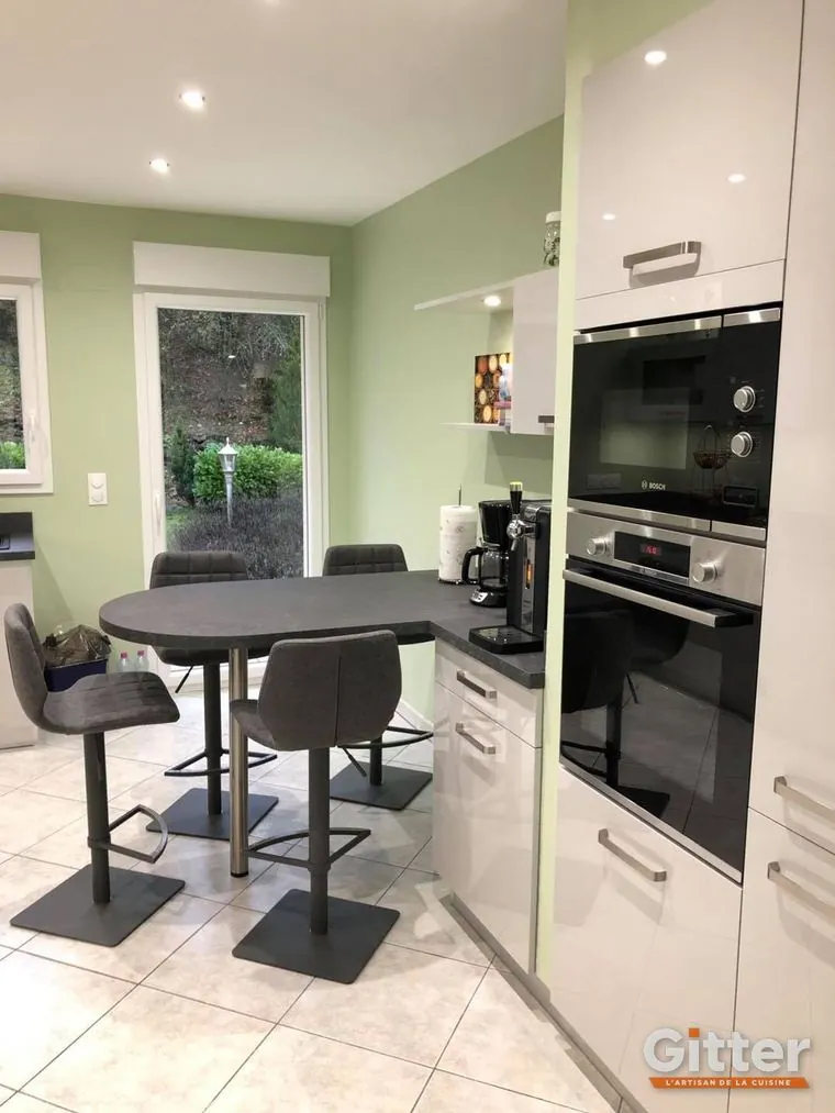 Cuisine ouverte, façades blanches brillantes, colonnes four et coin repas sur mur vert, réalisation Gitter à Rombas